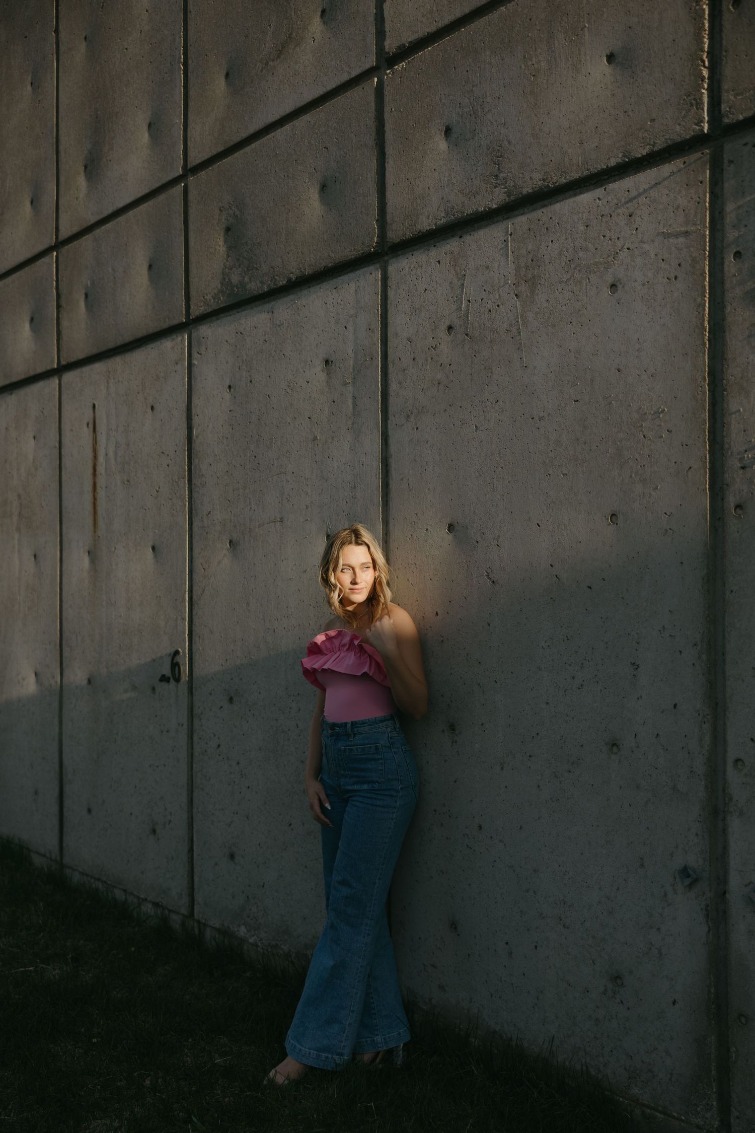 Artistic fashion photo with moody lighting showing pink top and flared jeans against industrial wall.