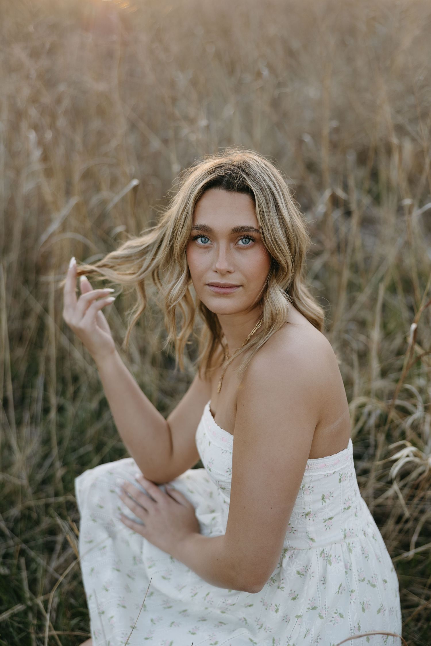 Romantic outdoor portrait in white floral dress amid golden dried grass at sunset.