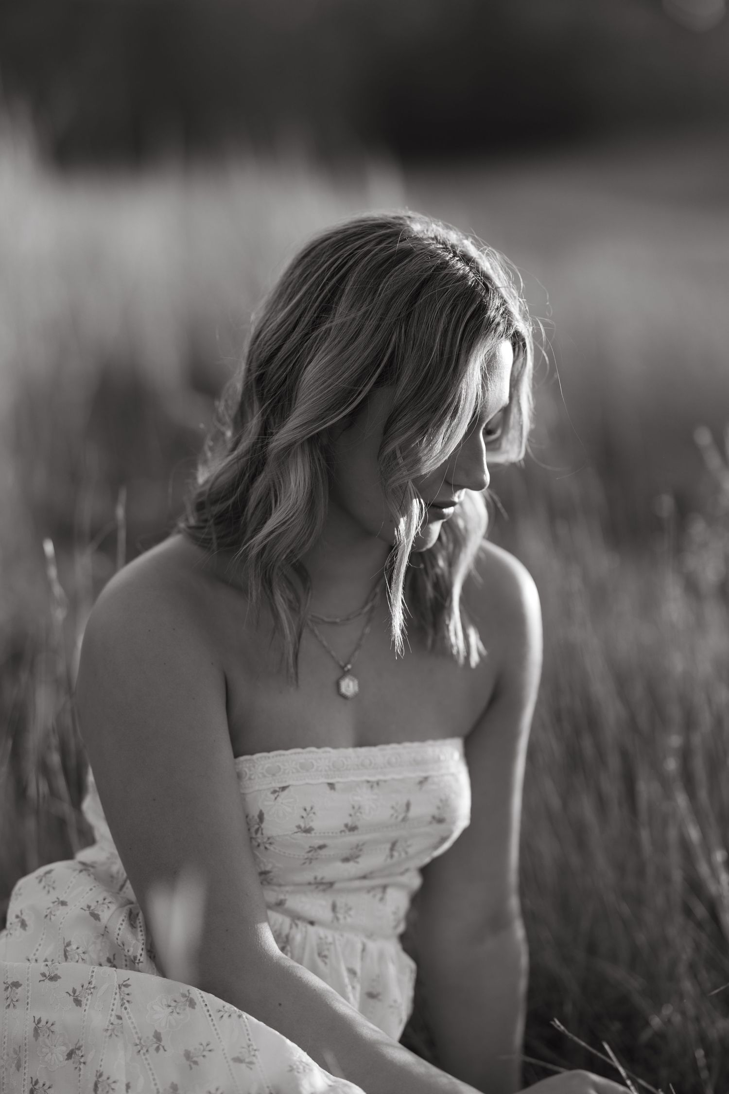 Artistic black and white photo of woman in floral dress sitting among tall grass.