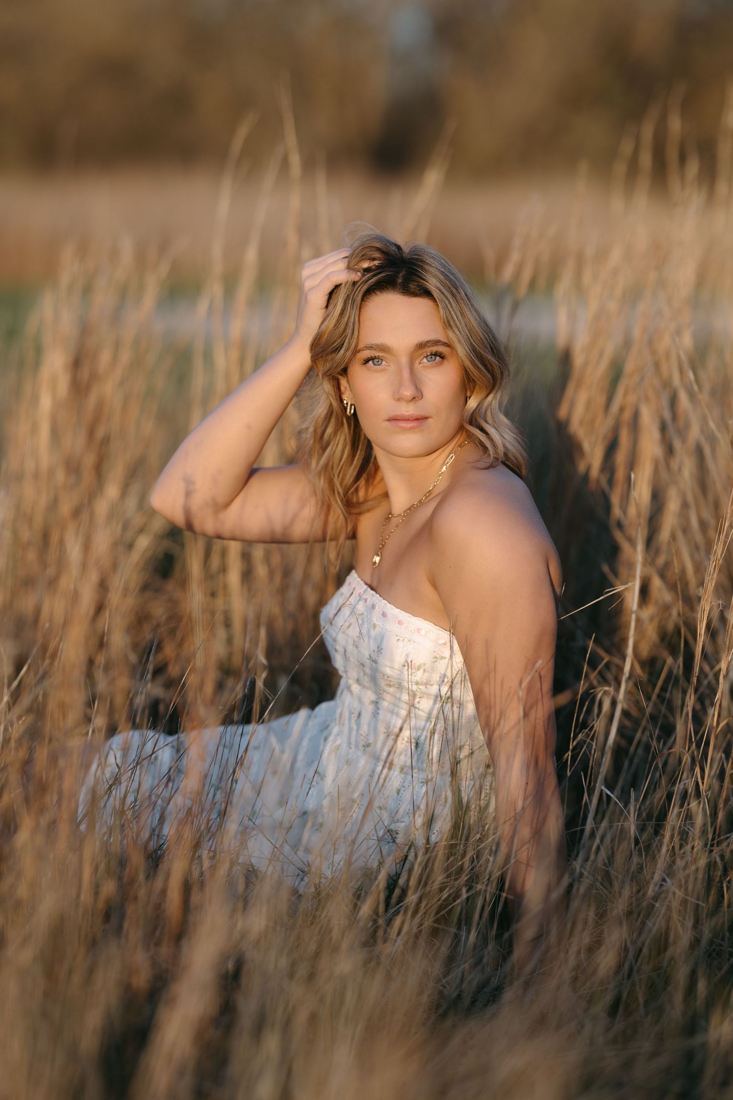 Lifestyle portrait of woman in white dress posing among tall grass during golden hour sunset.