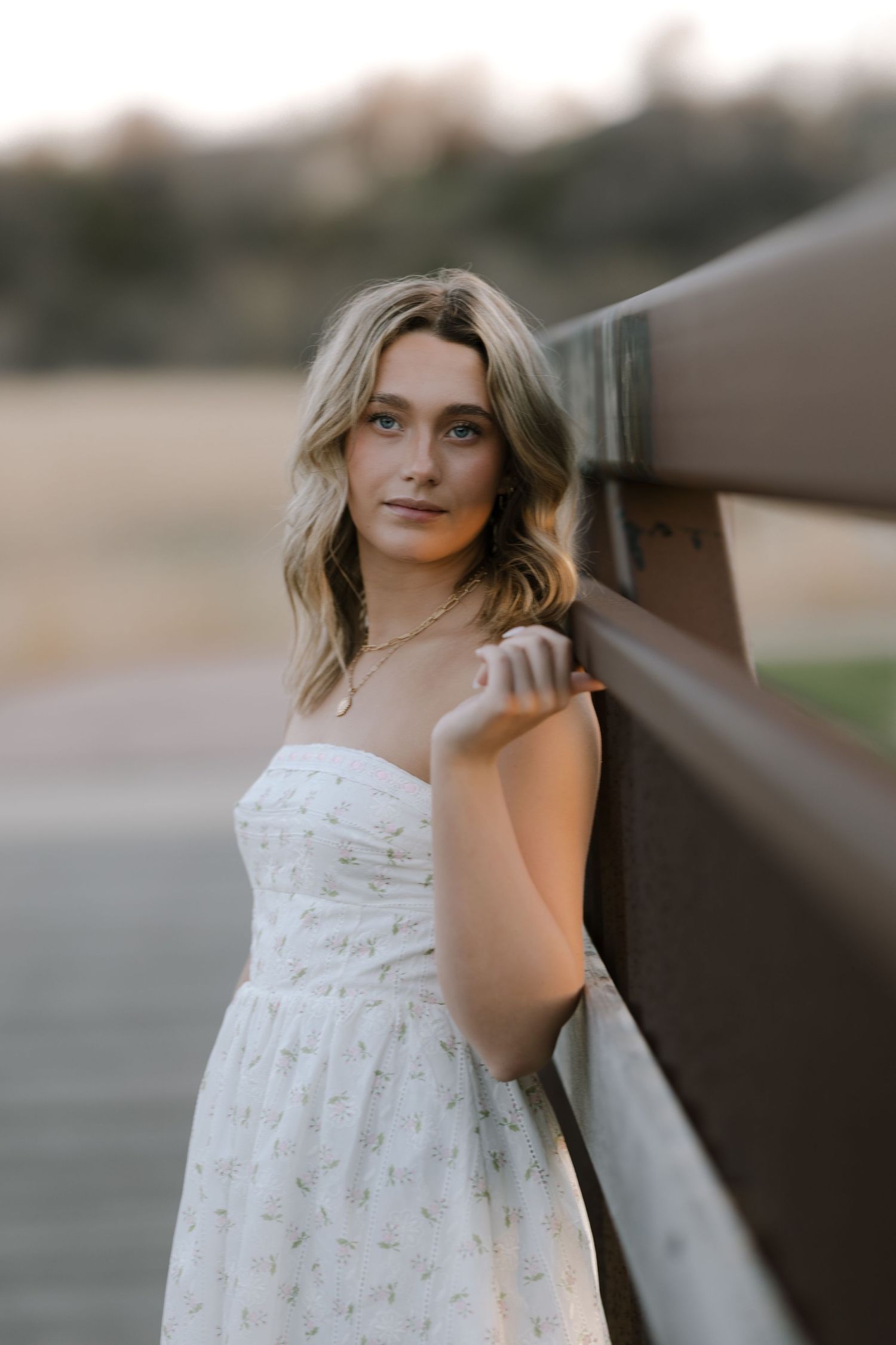 Portrait of woman in white summer dress leaning against wooden railing at sunset.