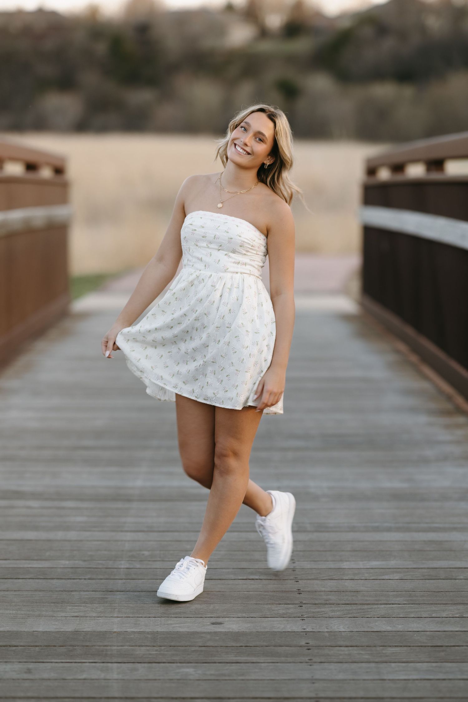 Candid portrait of woman in white sundress twirling on wooden boardwalk wearing white sneakers.