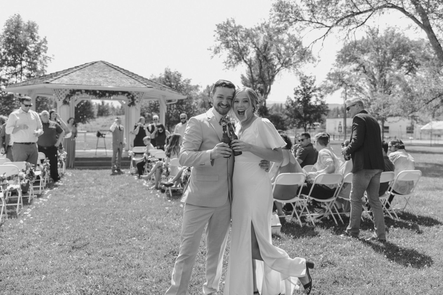 A black and white photo of a couple embracing outdoors at a casual event with folding chairs and guests in the background.
