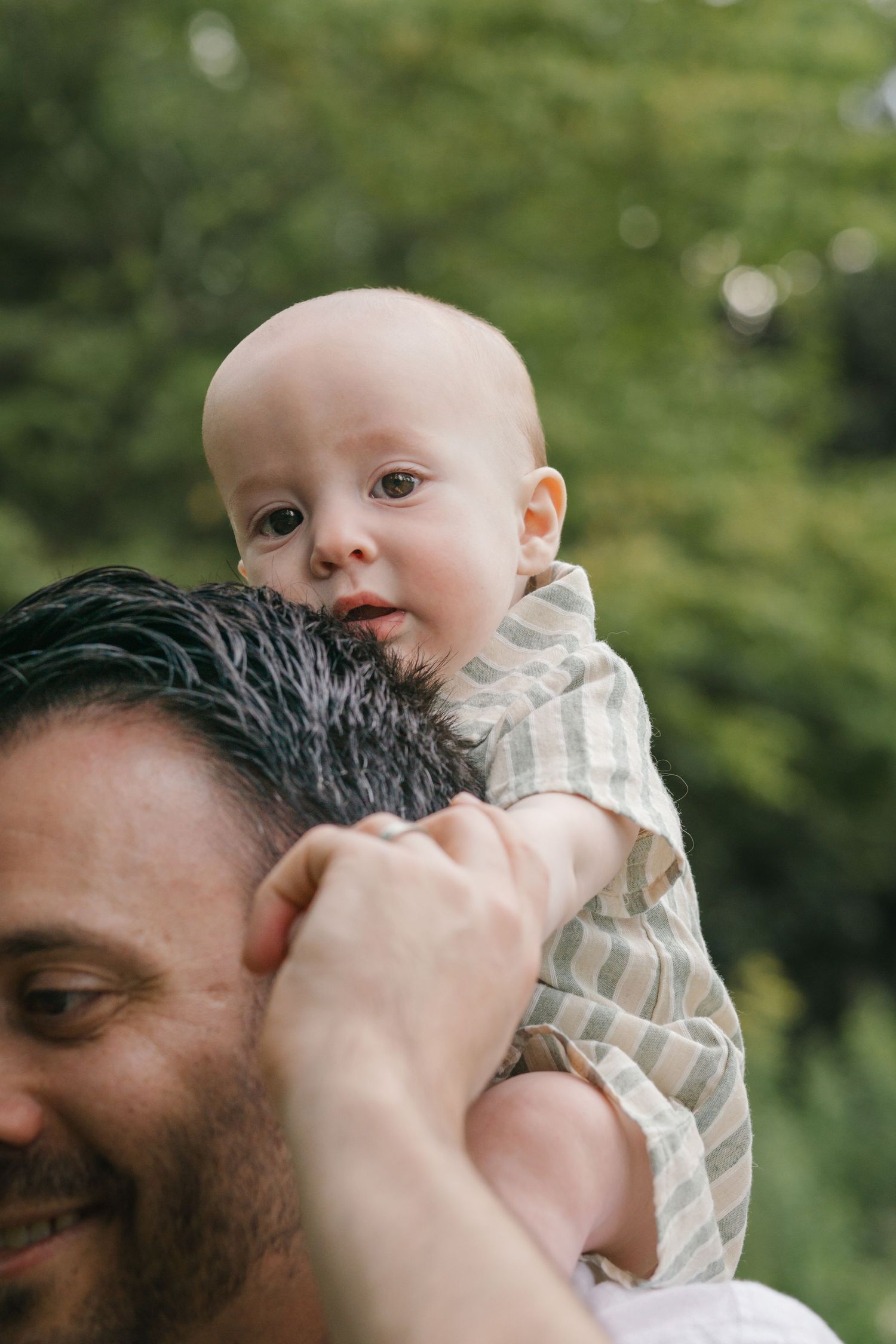 Close-up of a baby looking straight at the camera while resting on dad’s shoulder, surrounded by trees at Barclay Farmstead.