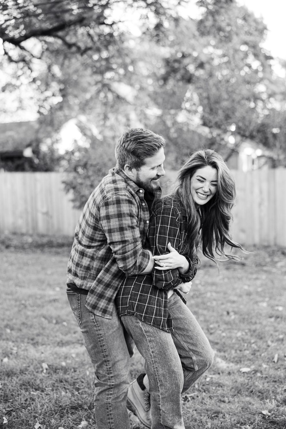 Black and white photo of a laughing couple embracing playfully outdoors in matching plaid shirts and jeans.