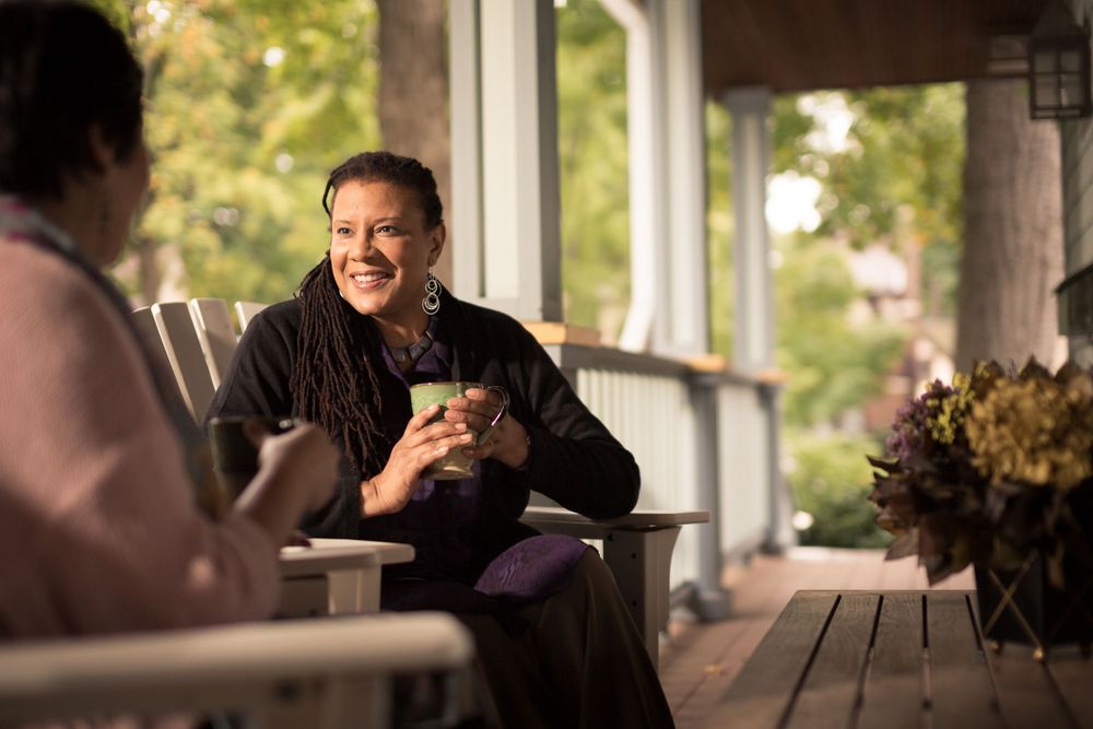 A casual conversation between people on a porch with warm sunlight and potted flowers.
