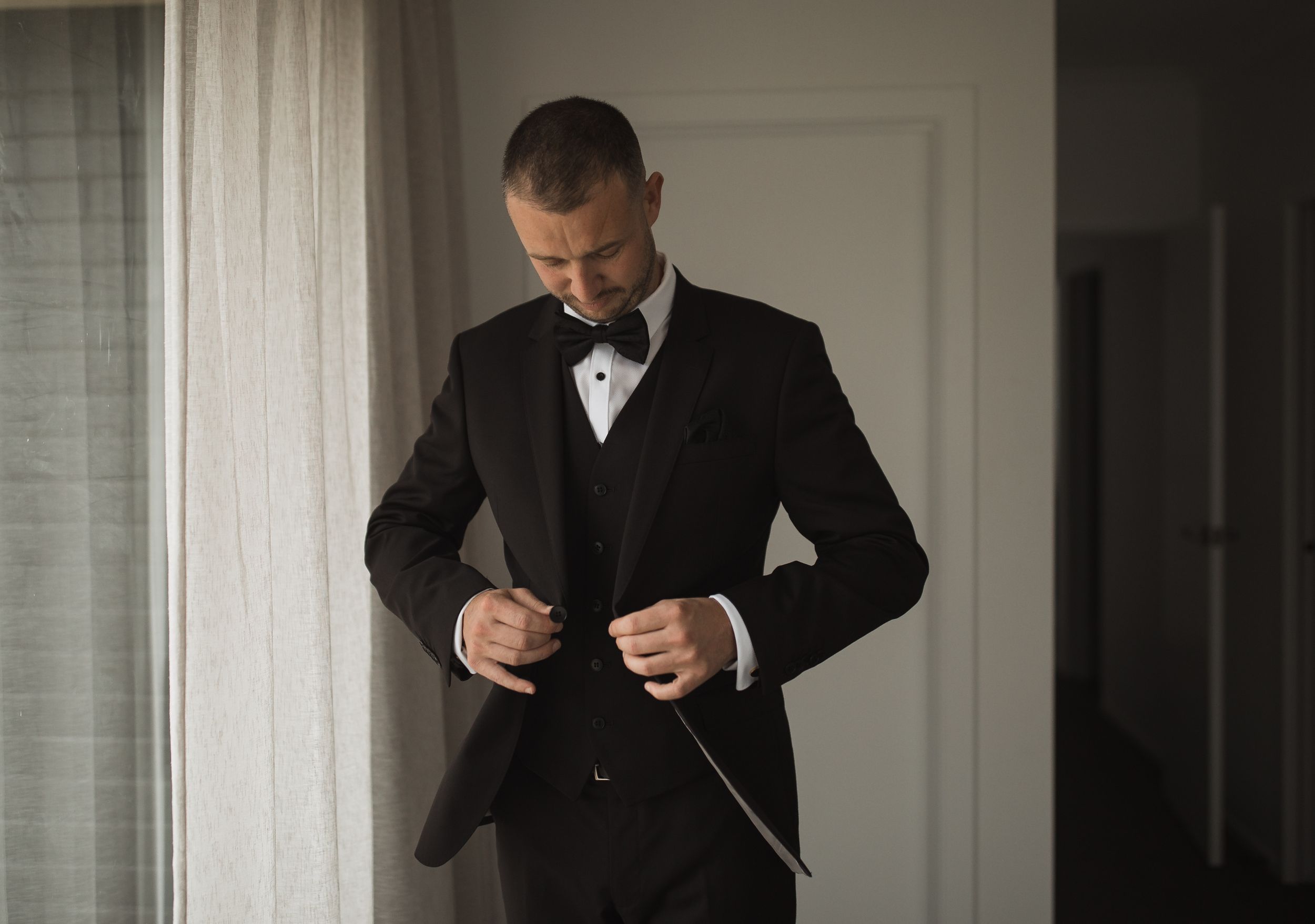 A groom adjusts his bowtie while getting ready in a minimalist room.