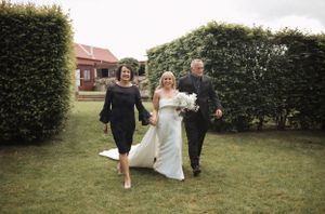 Wedding party running and laughing across a grassy field near rustic buildings.