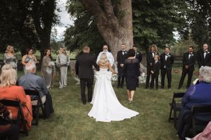 Outdoor wedding ceremony gathering under large trees with guests seated on white chairs.