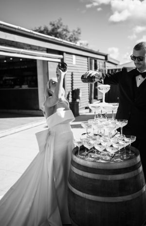 Champagne tower being set up on wooden barrel at outdoor wedding reception.