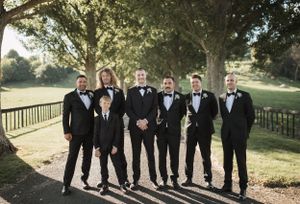 Groomsmen in black tuxedos pose together on a tree-lined path at an outdoor wedding venue.