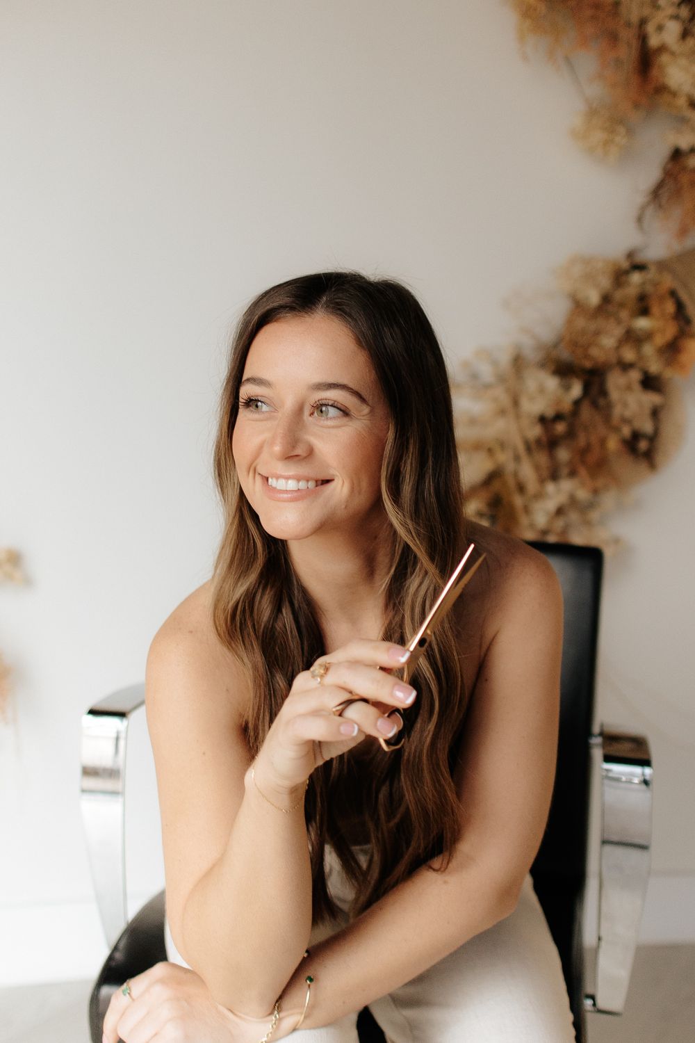 Woman with long brown hair smiling while holding chopsticks in natural lighting with a blurred background.