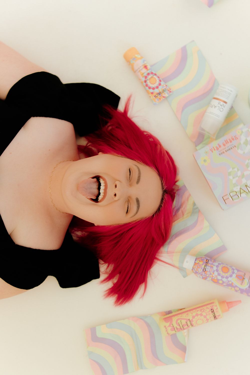 Vibrant red hair and joyful expression surrounded by colorful pastel beauty products on white background.