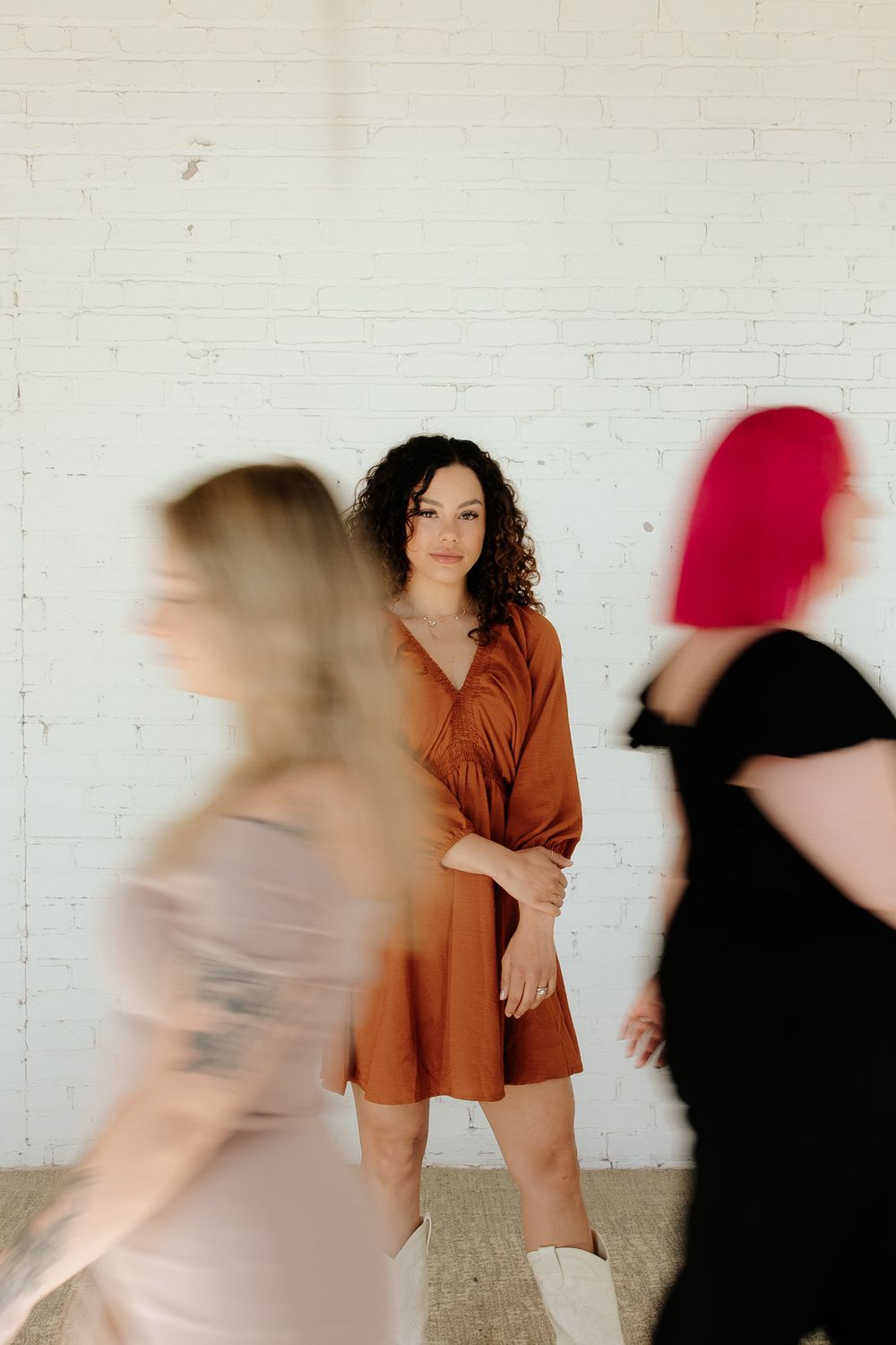 A person in a brown dress stands still while two others in motion create artistic motion blur against a white wall.