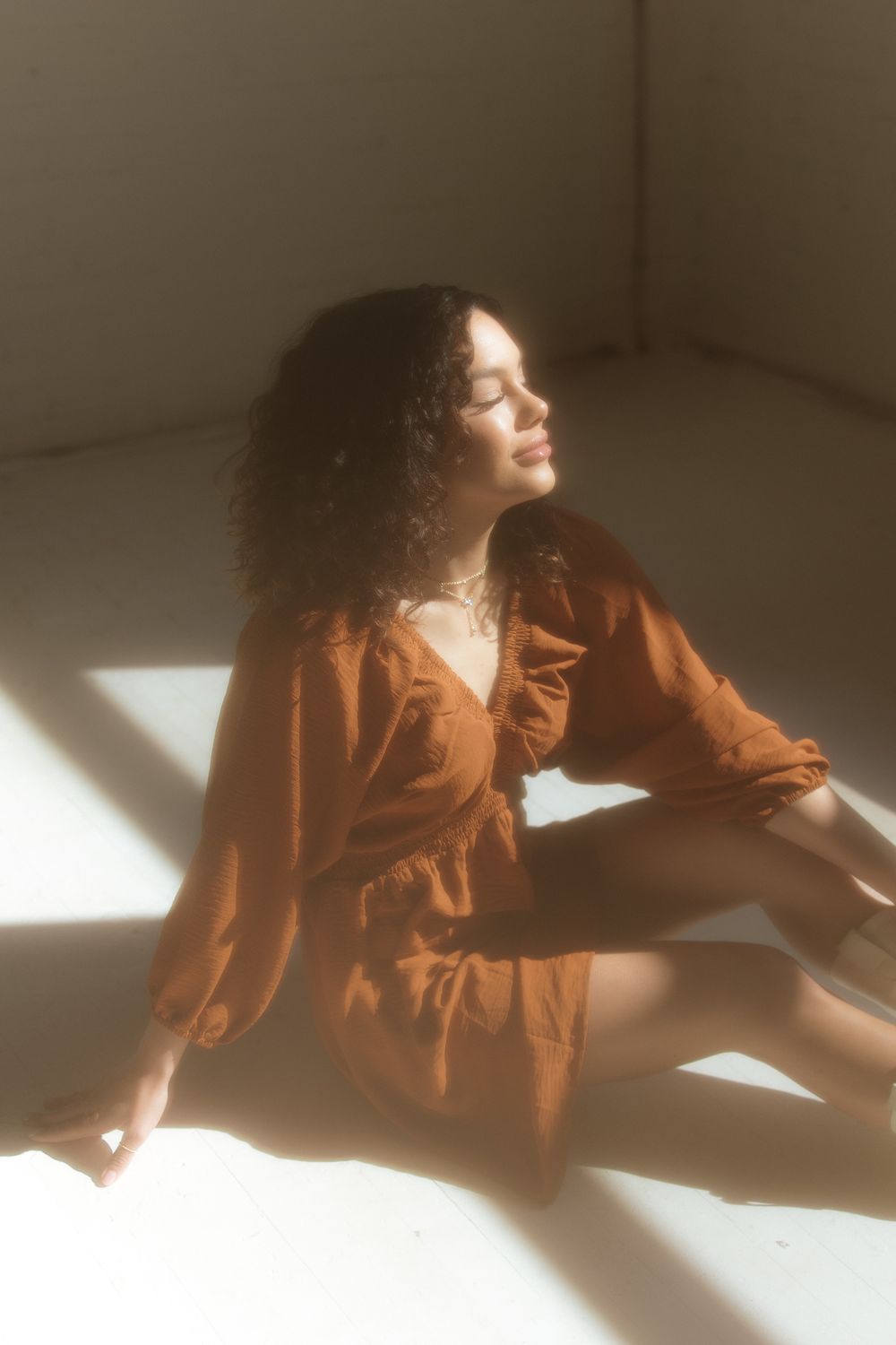 Dreamy portrait of woman in brown dress sitting in soft window light with curly hair creating ethereal mood.