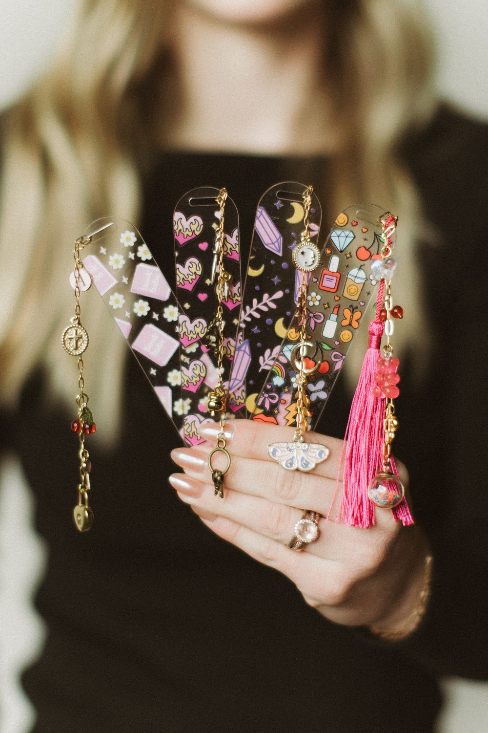 Hands holding clear phone cases decorated with colorful kawaii charms, hearts, and a pink tassel accessory.