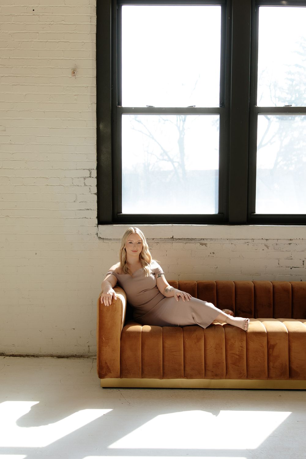A person in a beige dress lounges on a brown velvet sofa near a bright window.