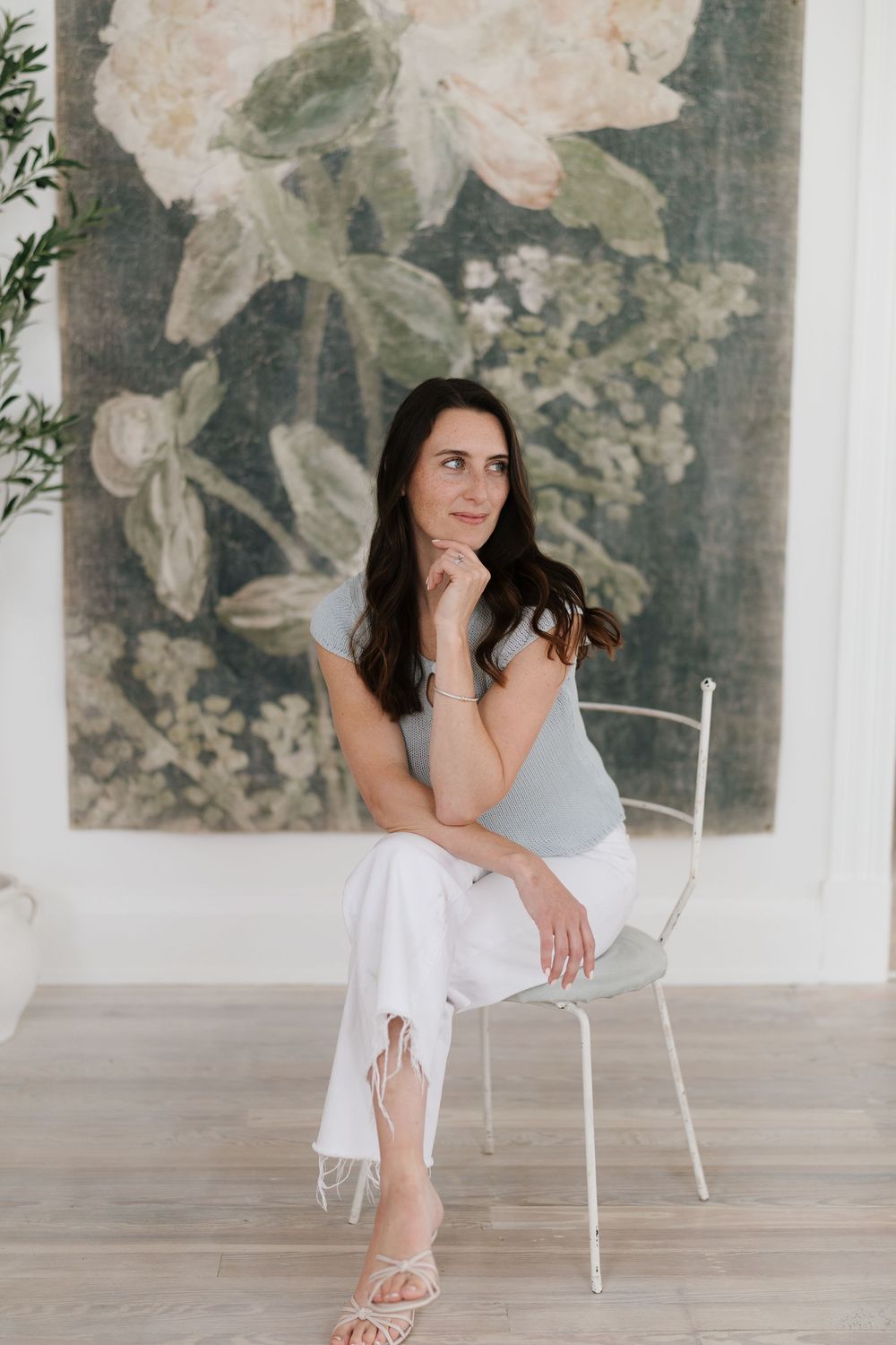 Woman in white pants and blue top sits thoughtfully in white chair before large floral wall art.