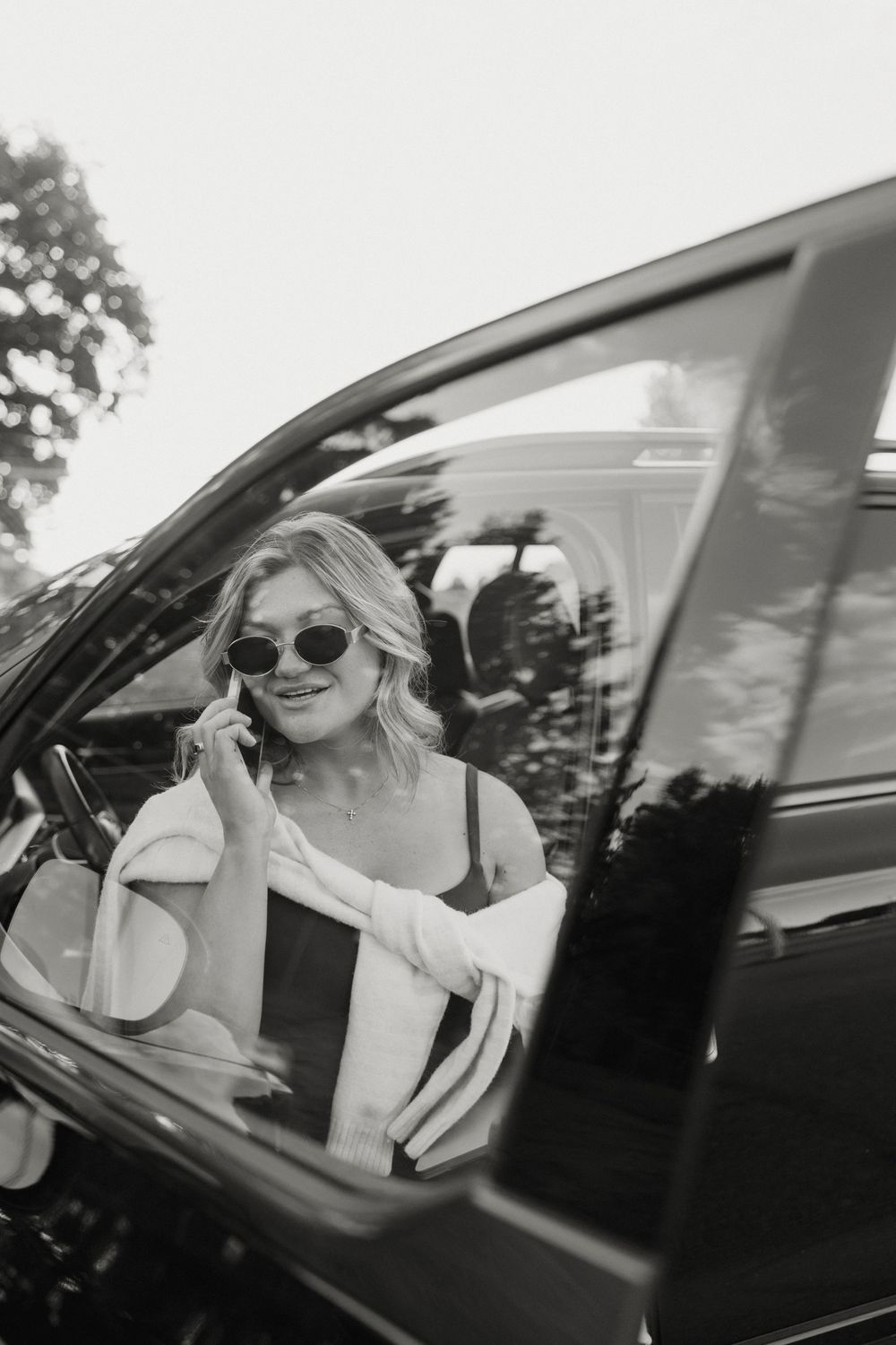 Black and white photo of a smiling blonde woman in sunglasses talking on phone while sitting in a car.
