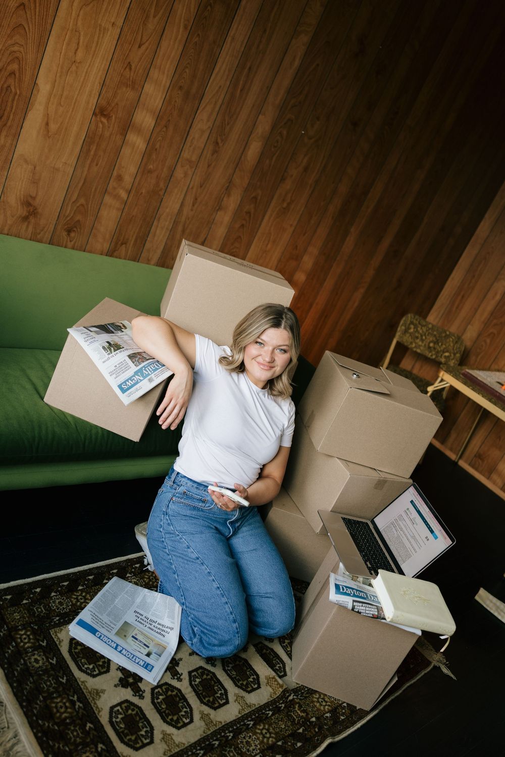 Woman sitting on floor surrounded by moving boxes and newspaper near a green sofa, smiling while holding a phone.