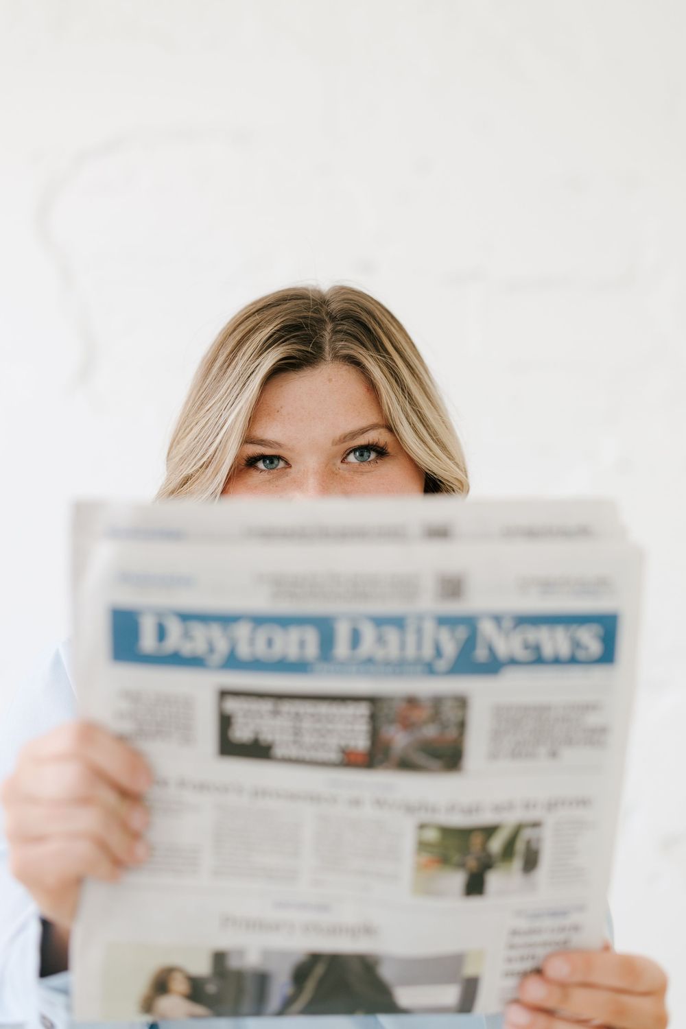 Woman peering over Dayton Daily News newspaper with curious blue eyes against white background.