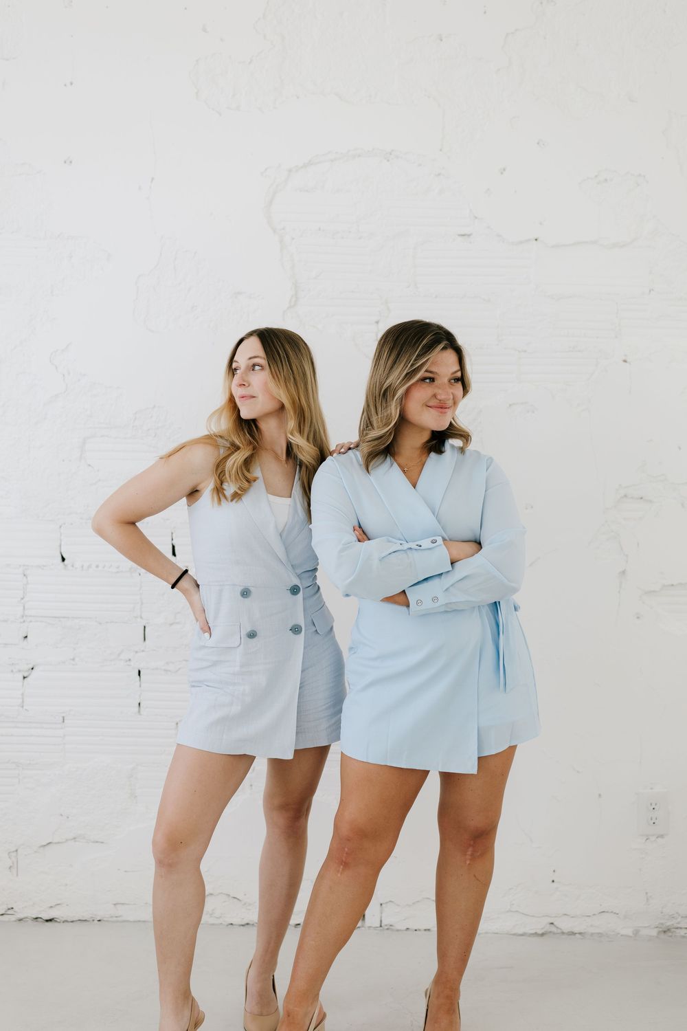 Two women in light blue outfits pose against a white brick wall, one in a dress and one in a blazer romper.