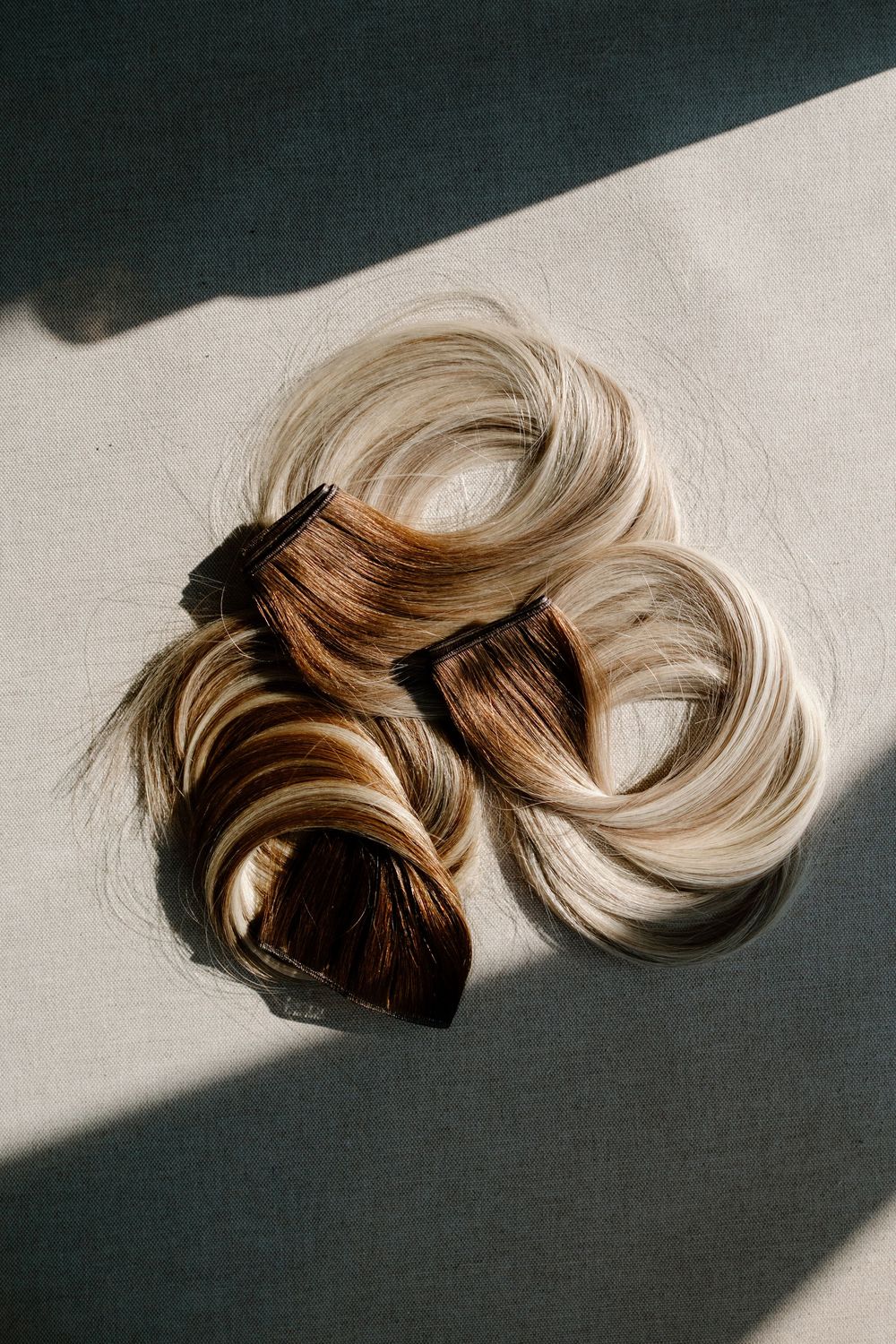 A blonde hair extension piece with dark roots lies in dramatic lighting against a white and black background.