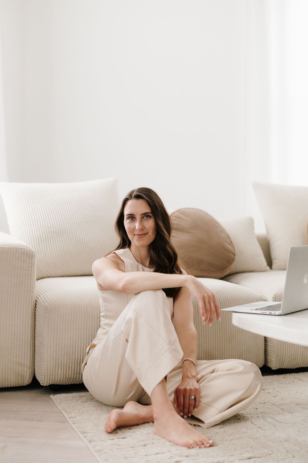 Woman in beige outfit sitting cross-legged on floor beside sofa with laptop, relaxed home setting.