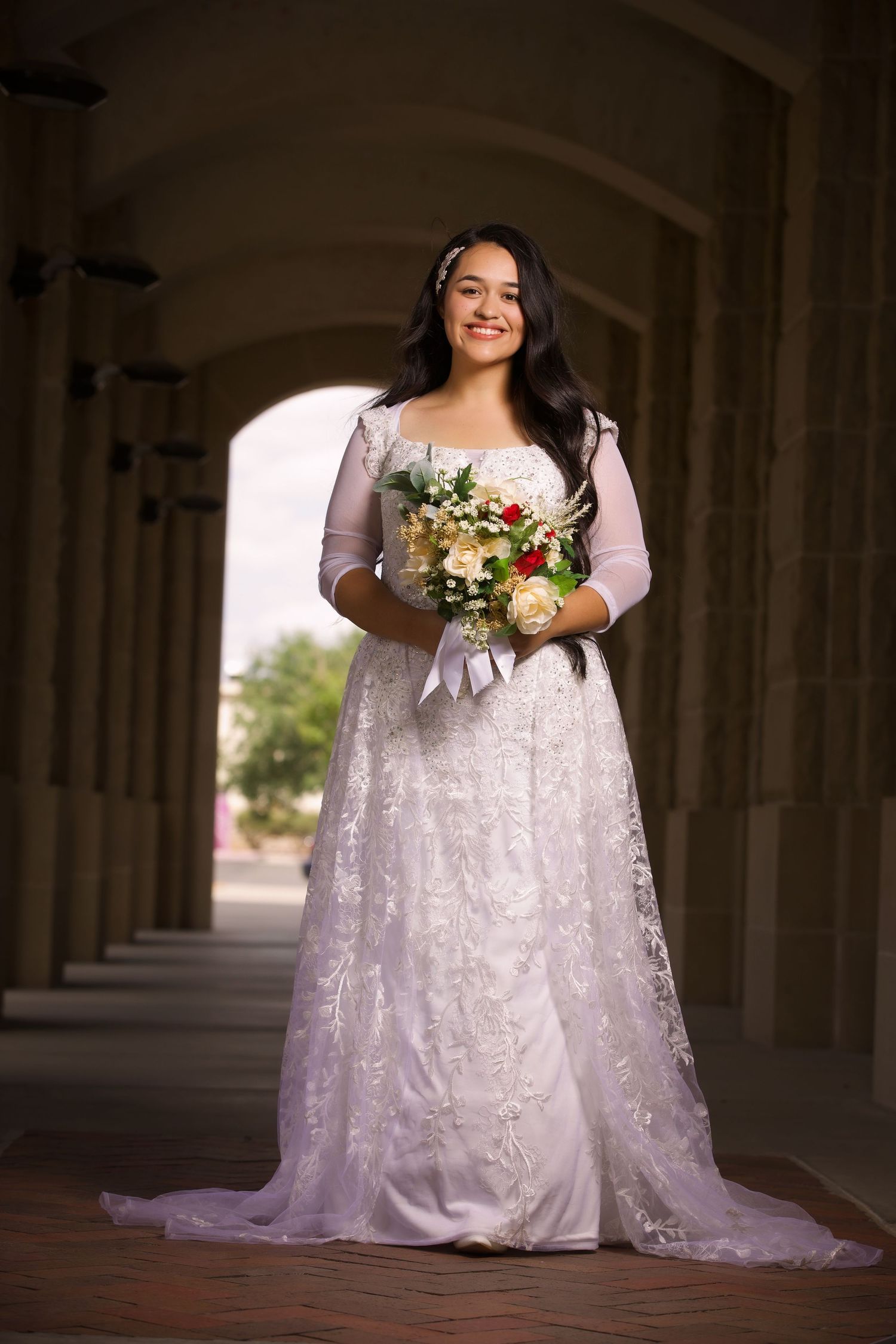 Beautiful bride in long white lace wedding gown with 3/4 sleeves poses with bouquet under stone archway.