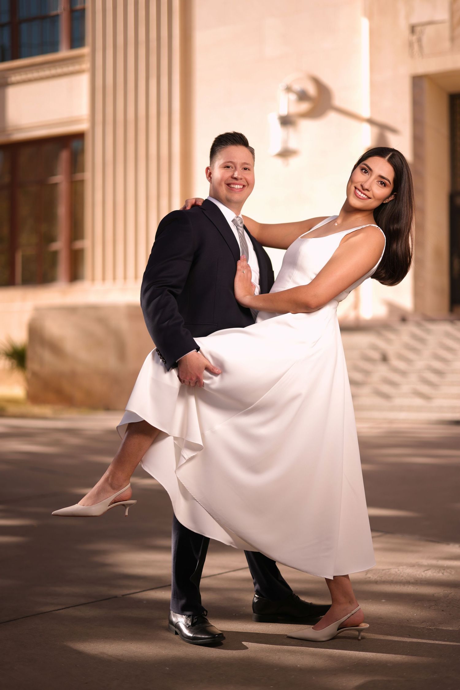 A couple in formal wedding attire share a romantic dance pose outside a building with columns at sunset.