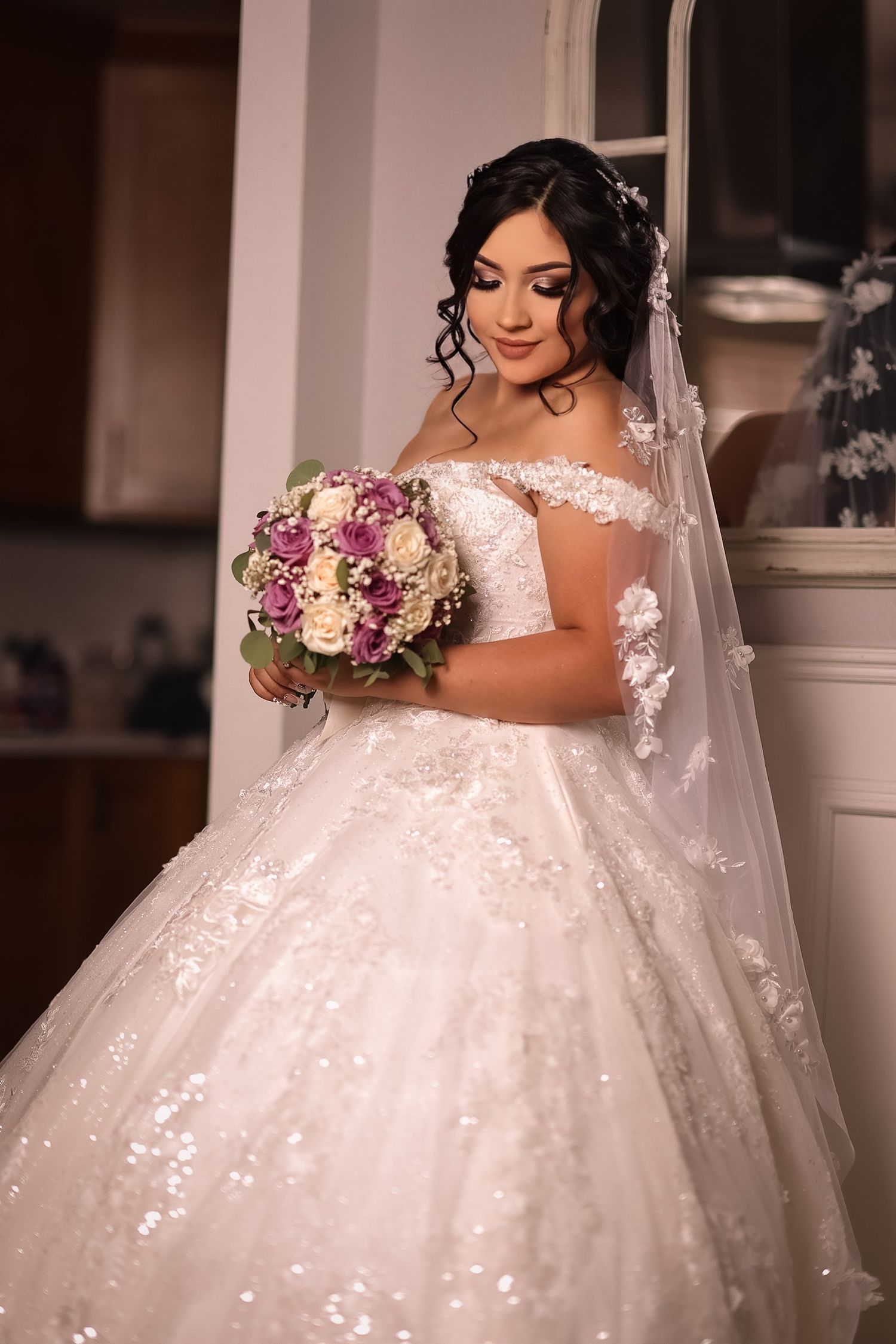 A bride in a sparkling white off-shoulder ballgown holds a pink and white bouquet while wearing a delicate lace veil.