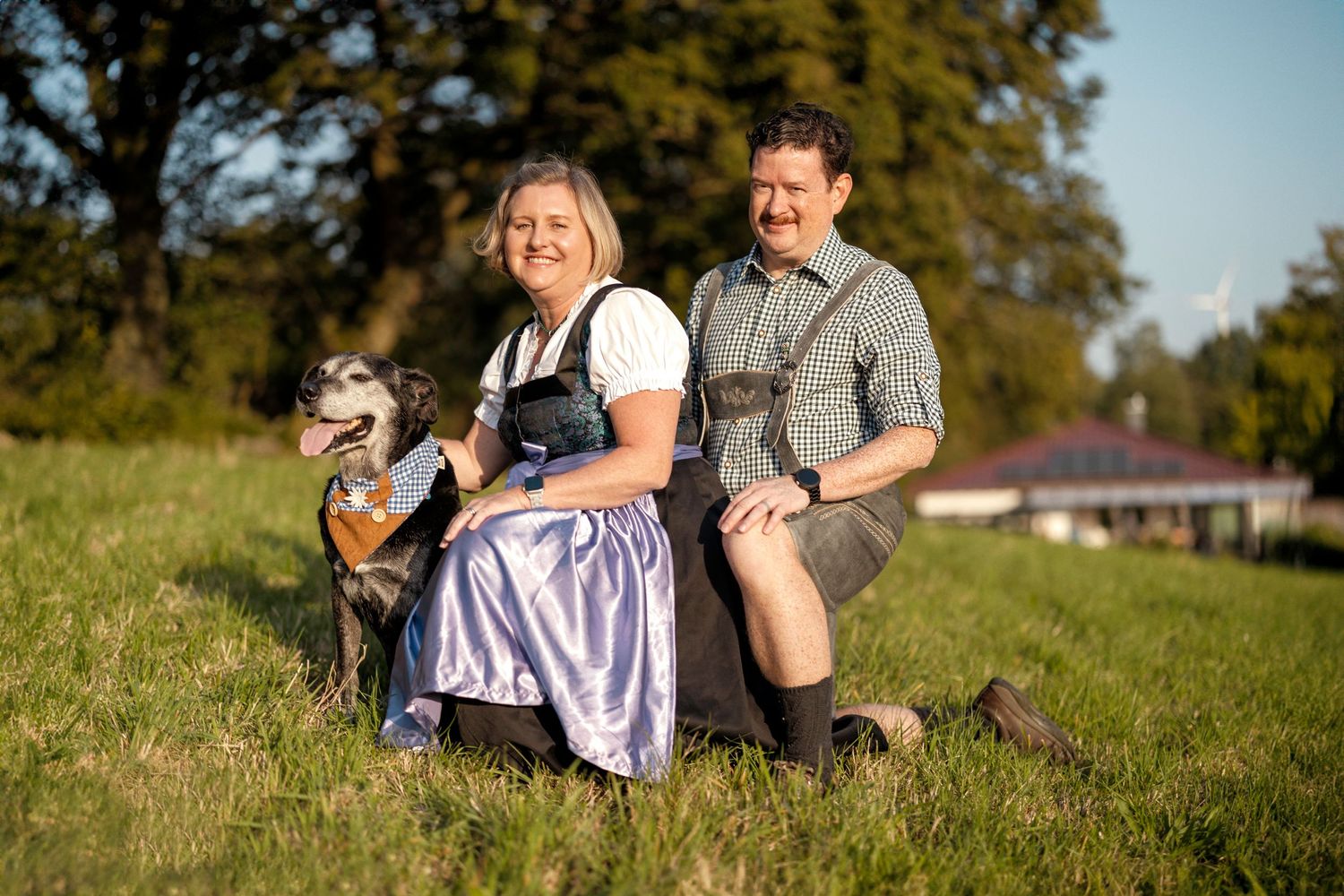 A couple in traditional Bavarian dirndl and lederhosen sits on a grassy hillside with their small dog in the sunlight.