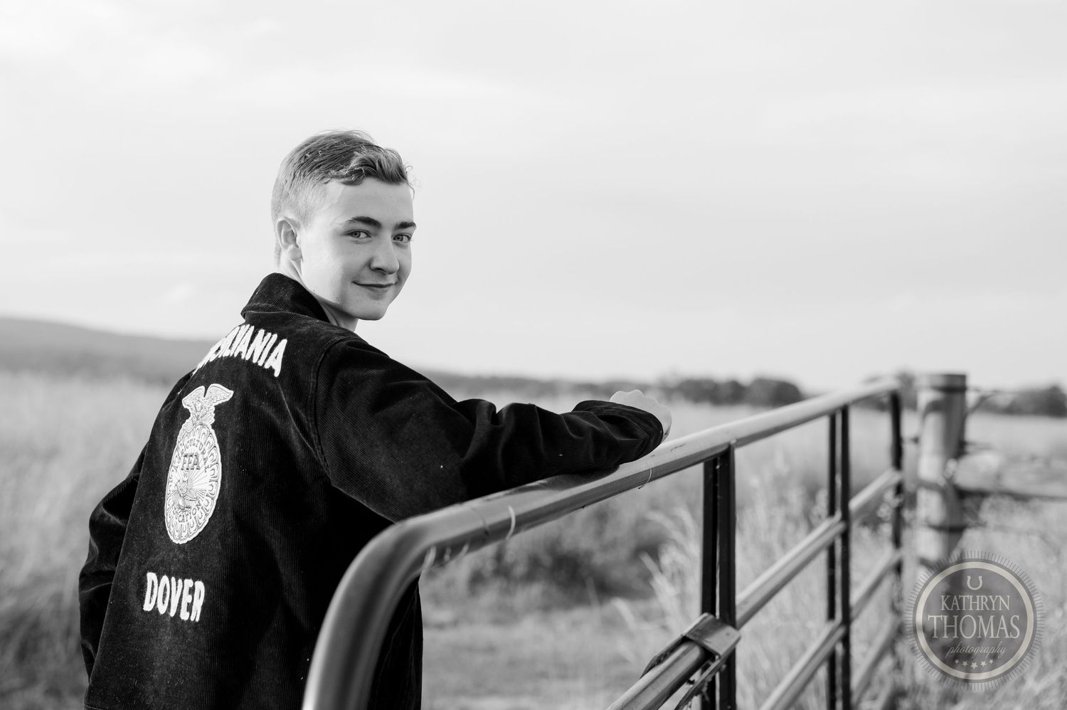 Black and white photo of student in FFA jacket leaning against metal railing in countryside.
