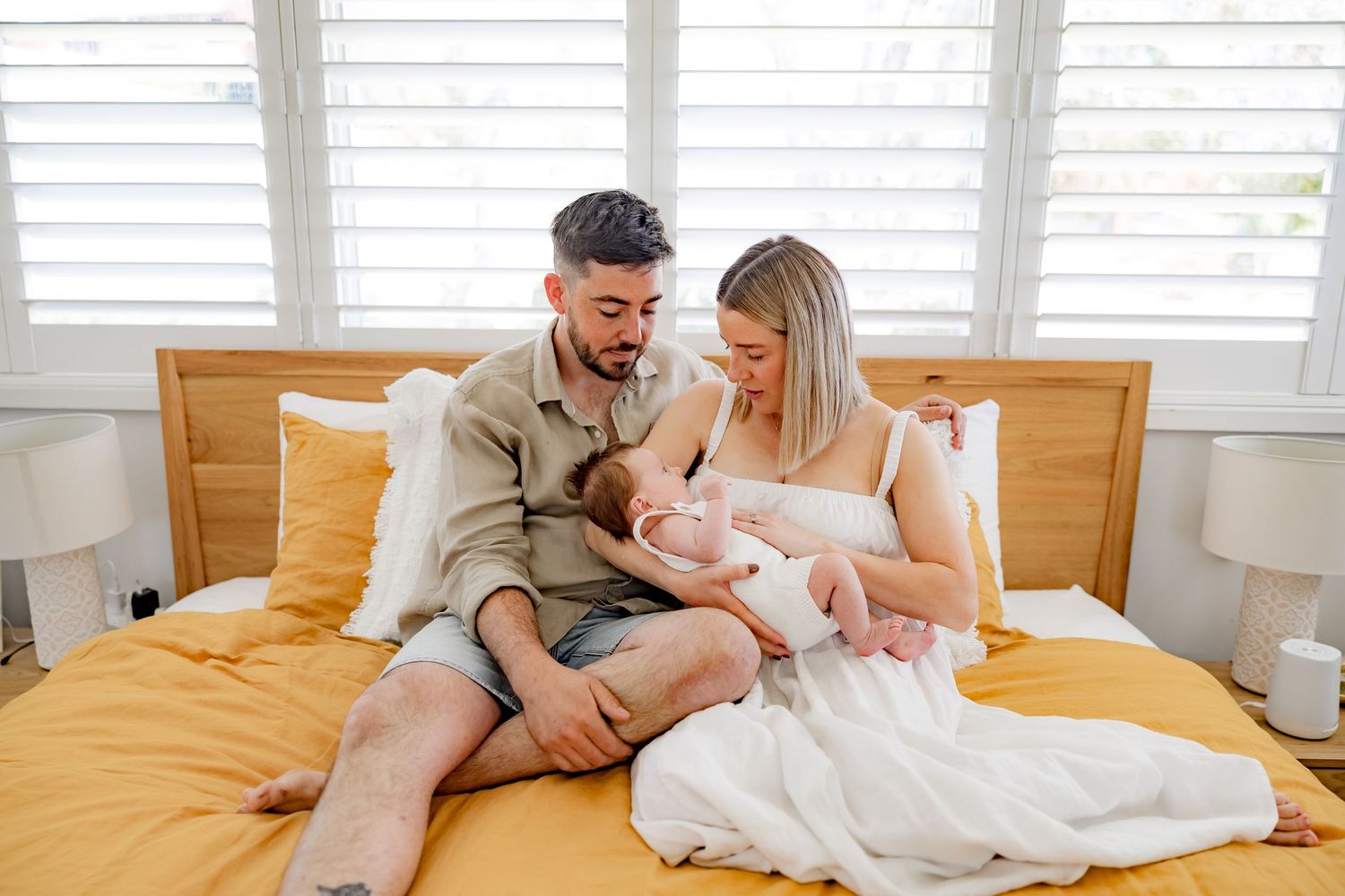 A family moment captured on a bed with yellow bedding and white shuttered windows.