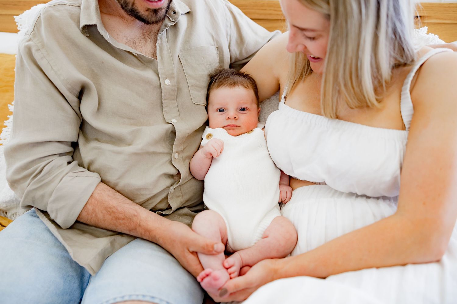 A cozy home scene with a small infant in white clothing sitting between two adults on a light colored sofa.