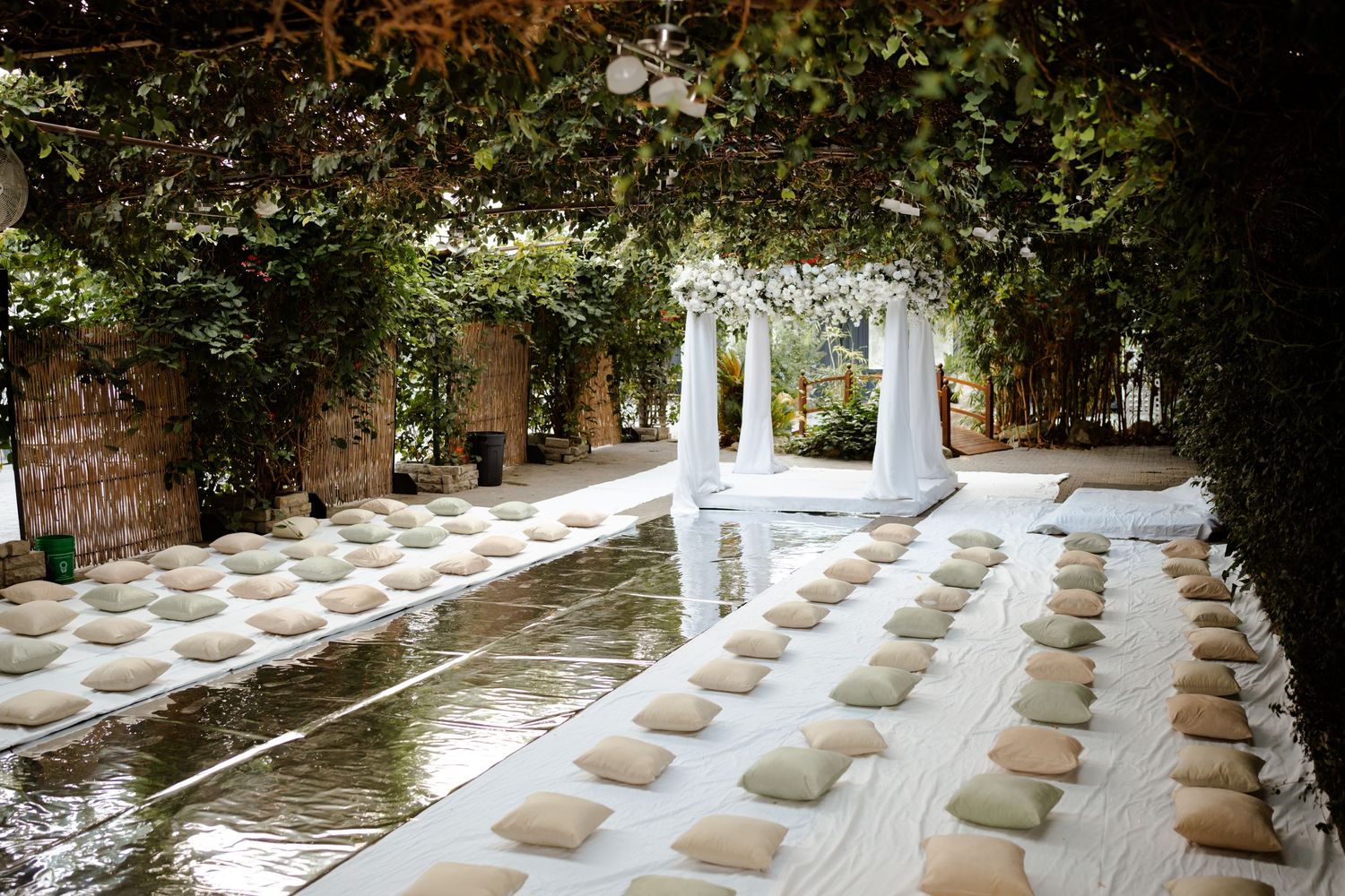 White folding chairs line an outdoor garden wedding ceremony setup with draped white fabric archway and lush greenery overhead.