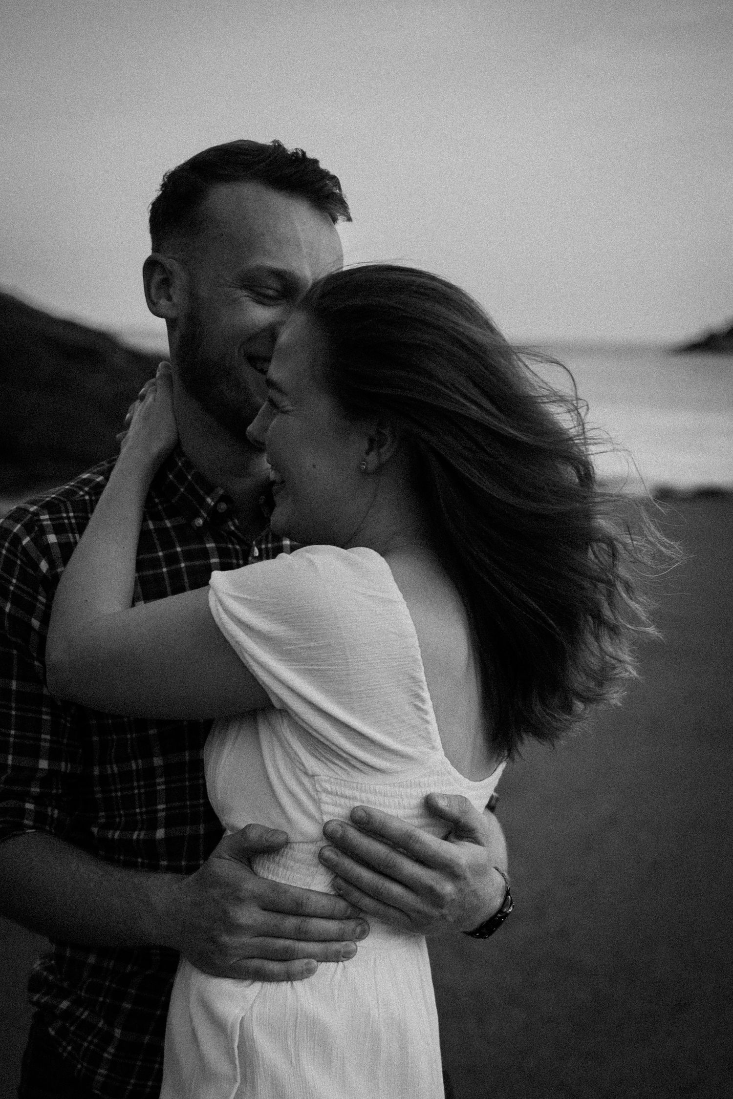 A black and white beach portrait captures a couple's intimate embrace with windswept hair.