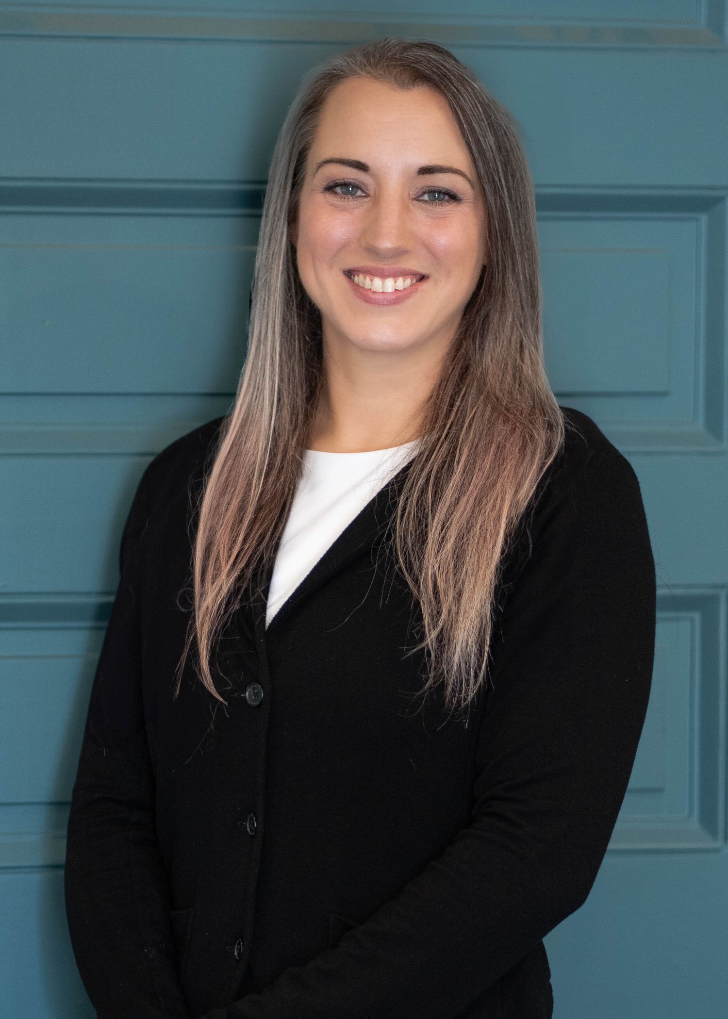 Professional headshot of someone with long brown hair wearing a black cardigan and white top against a teal door background.