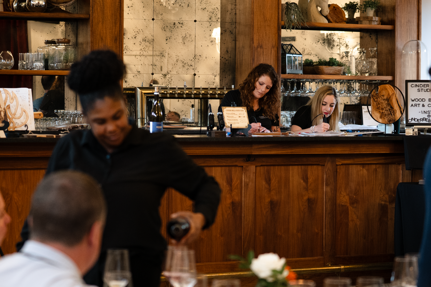 Restaurant staff serve customers from behind a rustic wooden bar with shelves of bottles and glassware displayed on the wall.