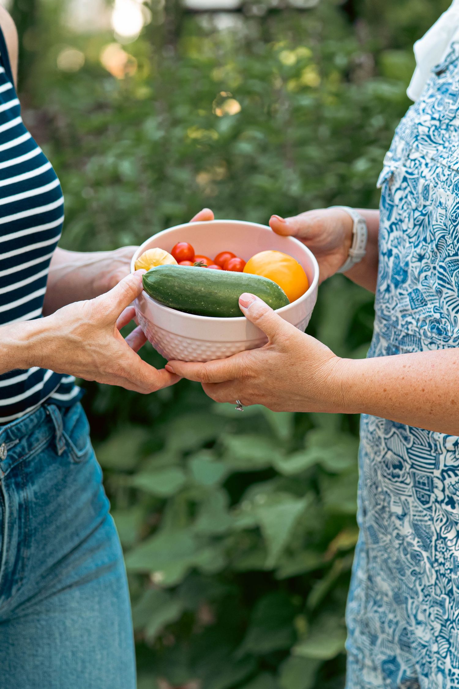 Two people passing a ceramic bowl filled with fresh vegetables and produce in an outdoor garden setting.