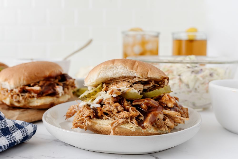 Pulled pork sandwiches with pickles and coleslaw on white plates against bright background.
