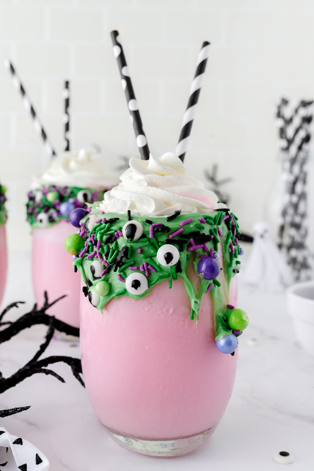 Halloween-themed milkshake with green drip, candy eyes, and black striped straws in a pink glass.