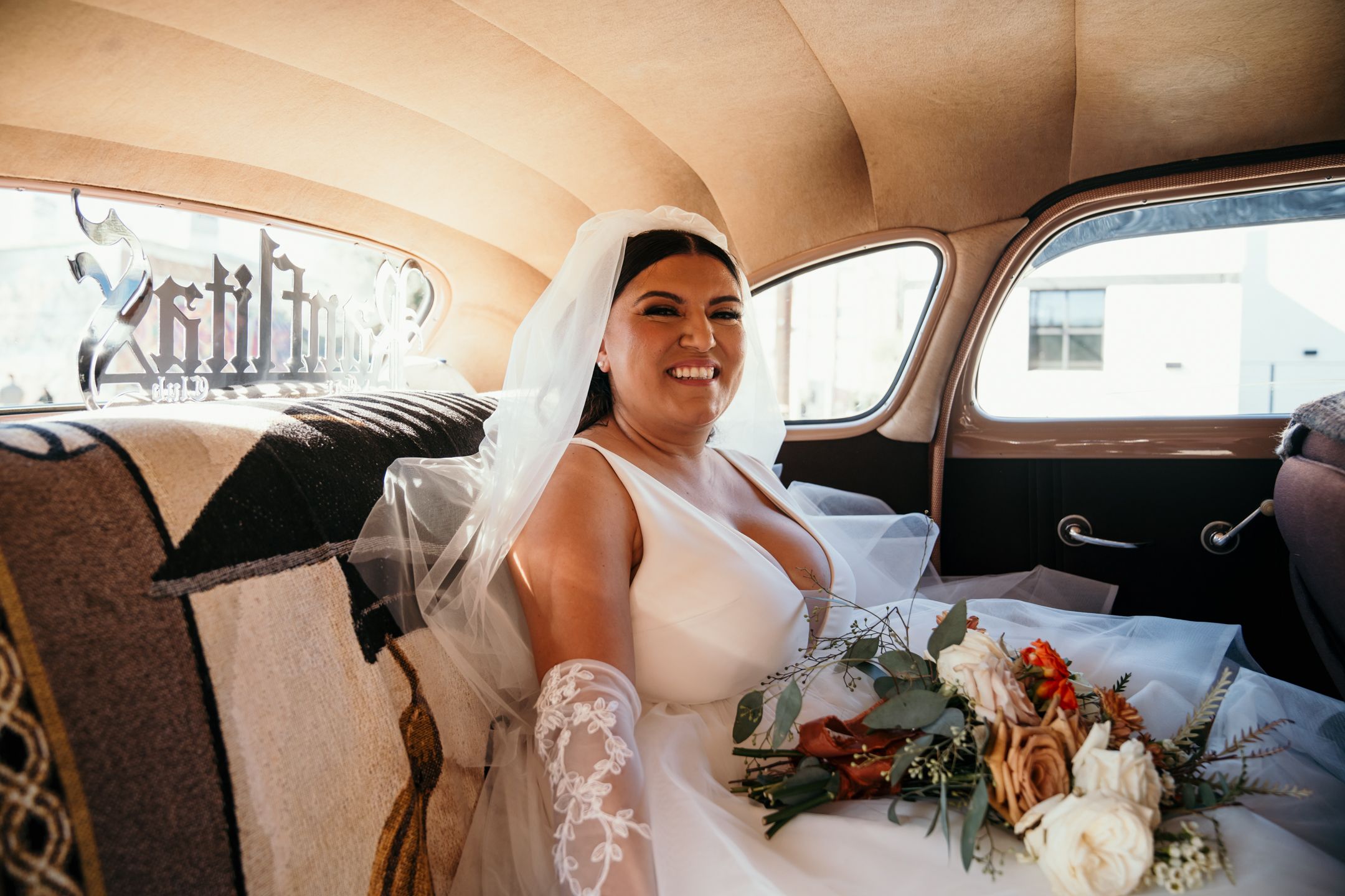 Person in white wedding dress sits inside vintage car interior.