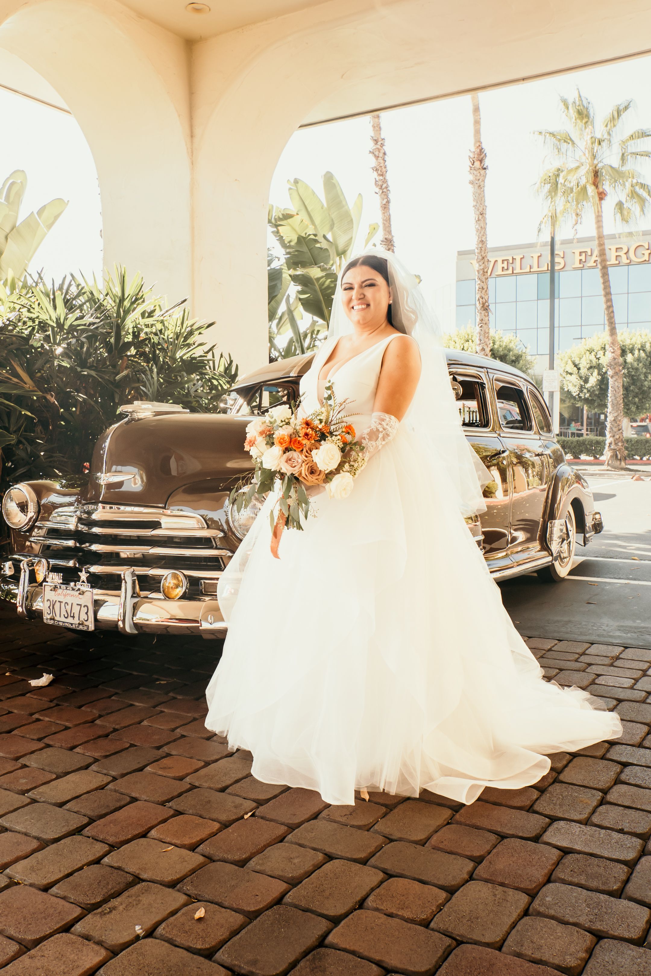 Person in white wedding gown holds bouquet next to classic car under palm trees.