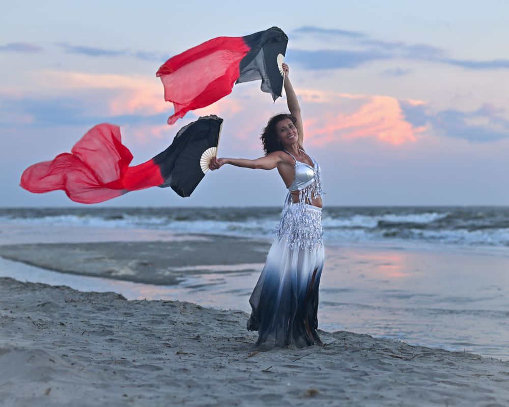 Dancer performs with red and black fabric against pastel sunset sky on coastal beach.