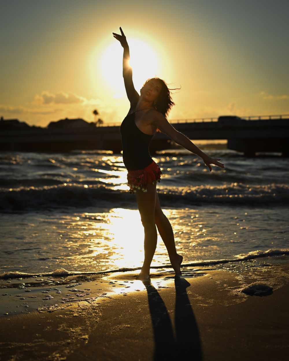 Silhouette of dancer poses gracefully on beach at sunset with waves in background.