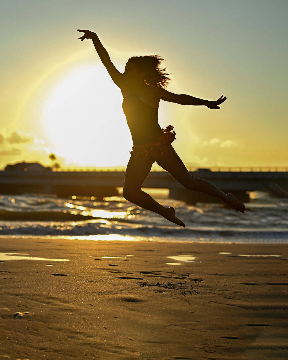 Silhouette leaps joyfully on beach with golden sunset and ocean waves in background.