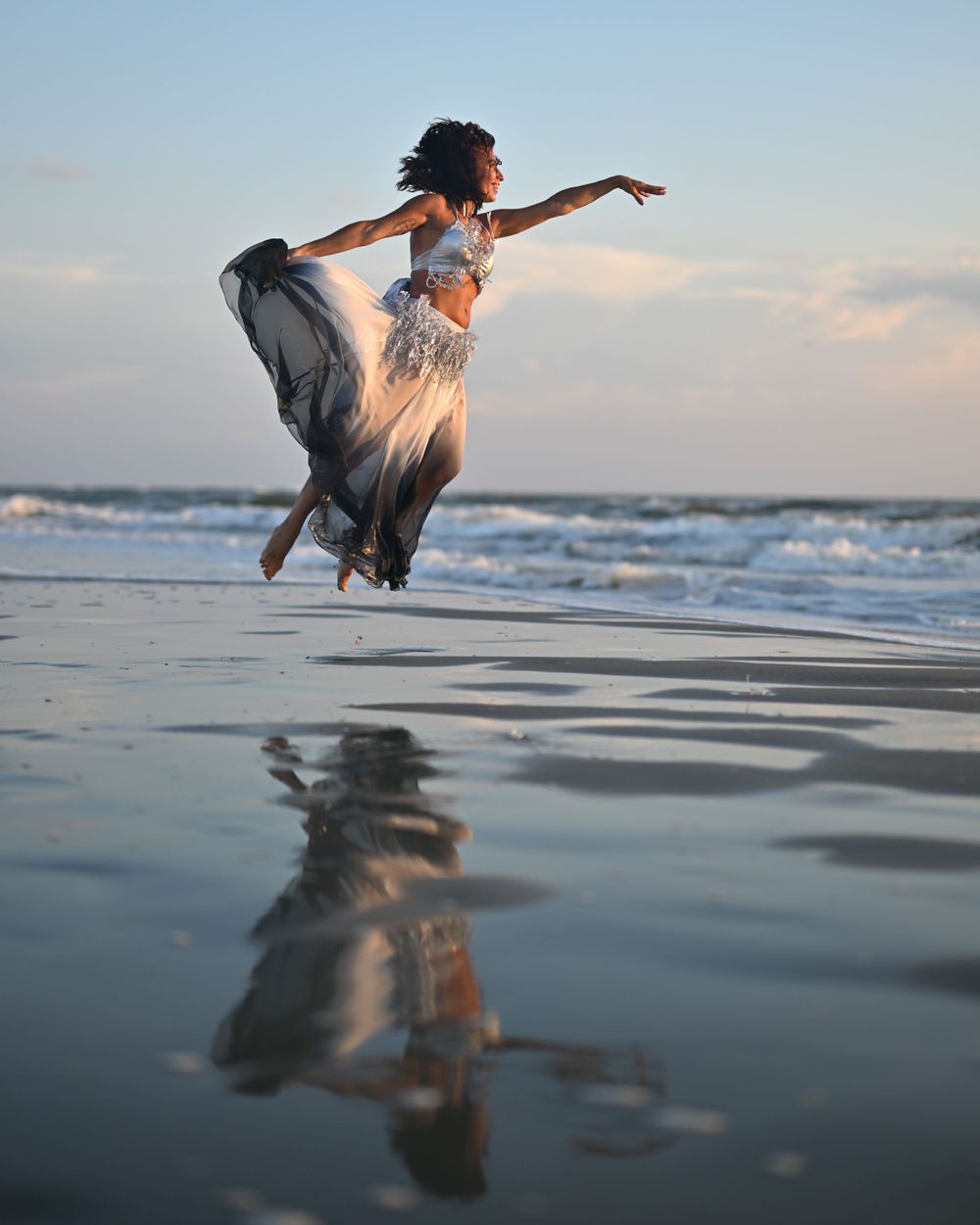 Dancer in flowing dress leaps gracefully over reflective beach waters at dusk.