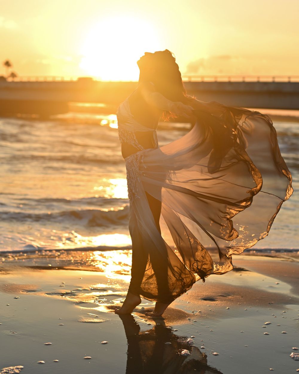 Figure in flowing dress dances on beach at sunset with waves and golden light.