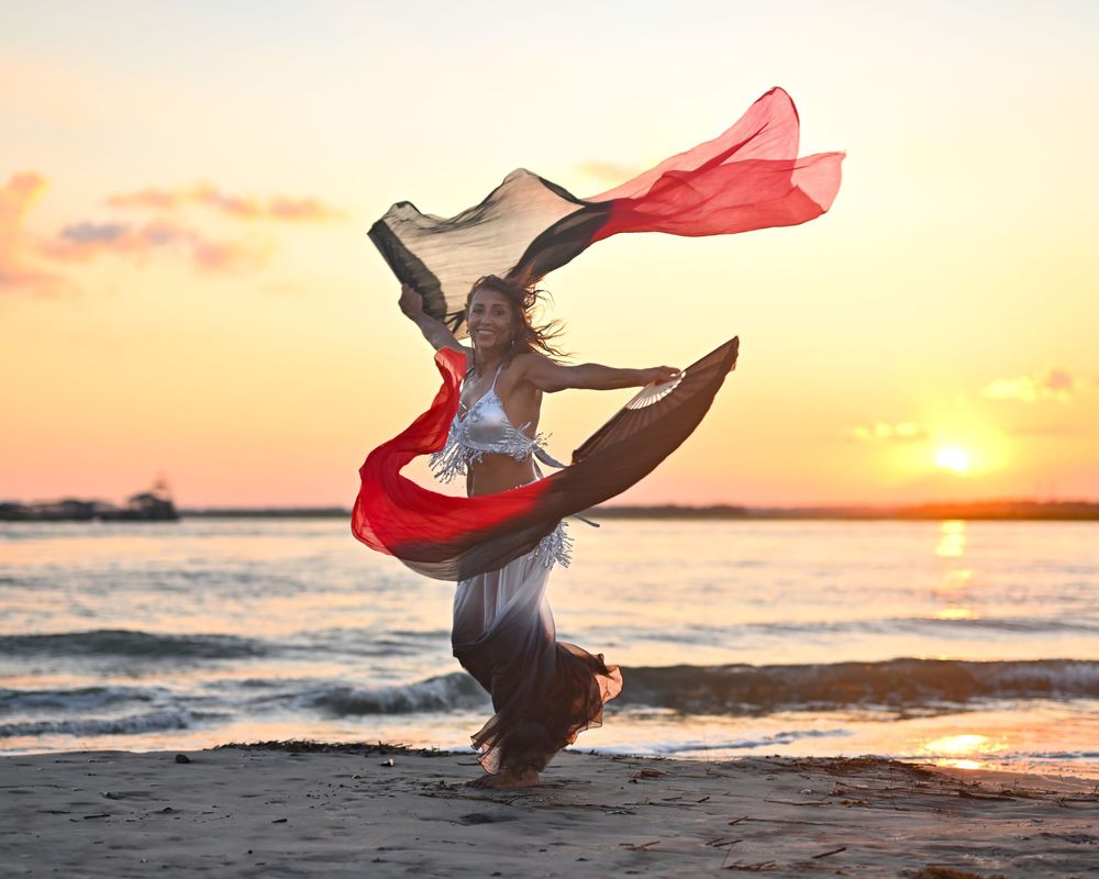 Dancer leaps with flowing red fabric against dramatic beach sunset.
